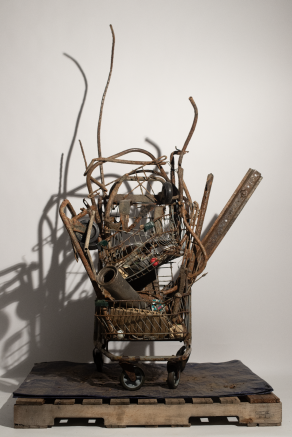 Sculpture made out of rusty scrap metal, assembled in an old shopping cart, displayed on a pallet in front of a white wall. 