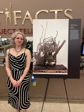 Woman stands next to a poster image of a sculpture made out of rusty scrap metal, assembled in an old shopping cart, displayed on a pallet in front of a white wall. 