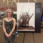 Woman stands next to a poster image of a sculpture made out of rusty scrap metal, assembled in an old shopping cart, displayed on a pallet in front of a white wall. 