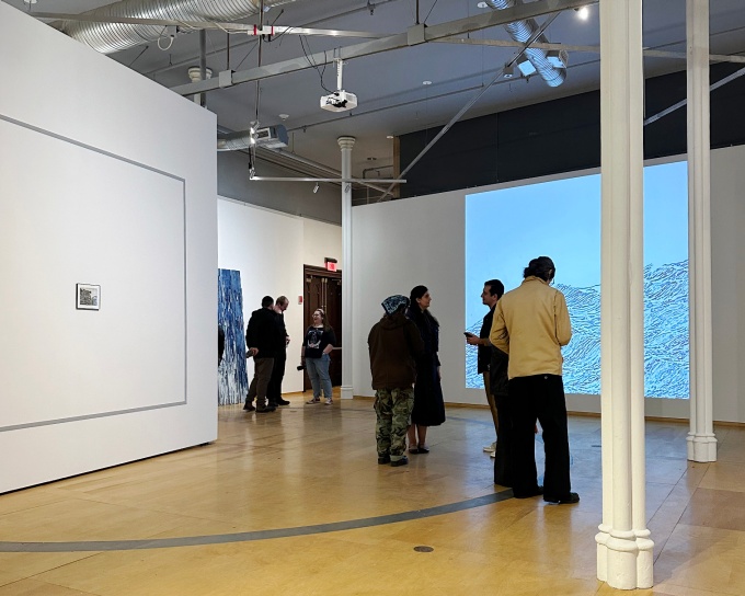 A handful of people stand in a gallery with wood floors, white walls, and a water-like artwork projection on far wall. 
