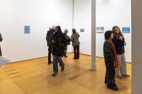 Zoom image: people standing in an art gallery with white walls and wood floors, and shades of blue artworks on the walls