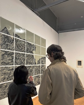 Two people facing a wall of art panels made on glass rectangles with wave patterns, hung in a grid on white walls. 