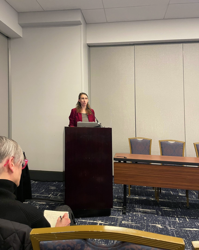 Zoom image: A woman standing at a podium in a hotel conferenence room, with an audience memebr taking notes in the front row.