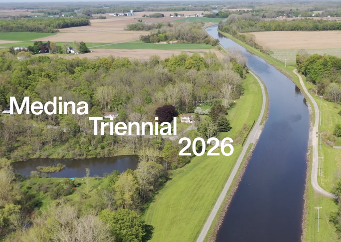 Zoom image: birds eye view of forests and farmlands bordering a canal, with "Medina Triennial 2026" overlaid in white text