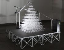 A tower of champagne glasses on a scaffolding platform a couple feet off the ground, with a currency vending machine and mechanical arms. 