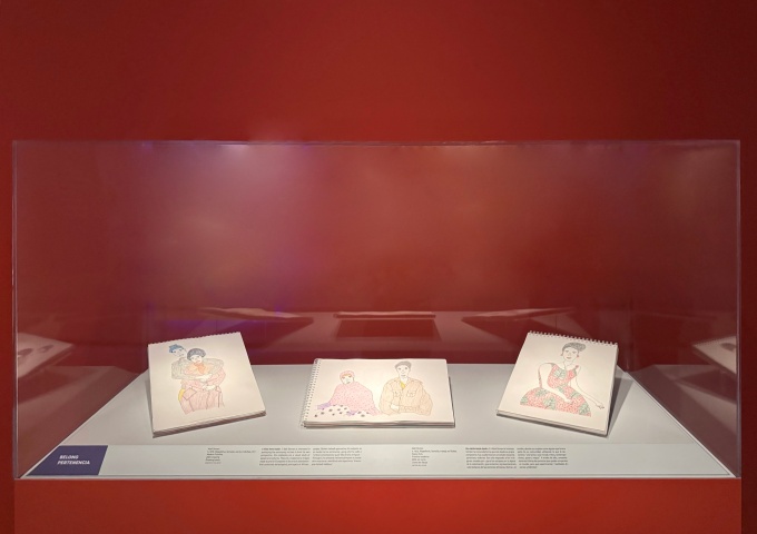 Zoom image: Vitrine containing three open notebooks aof drawings of people, in front of a red wall.