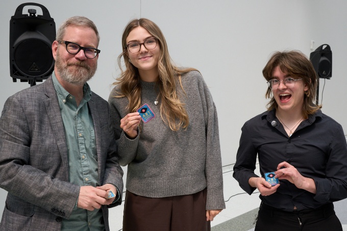 Three people stand in front of a white wall and hold up embroidered patches. 