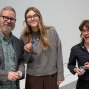 Three people stand in front of a white wall and hold up embroidered patches. 