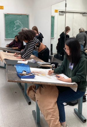 A line of students sketching during Figure Drawing Class, at angled drawing tables, with miscellaneous drawing implements and various types of paper on their work stations in front of them, including colored pencils, charcoal, and crayons. 