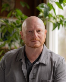 Headshot of artist Michael Bosworth, wearing a gray button up shirt, in front of a plant and window. Bosworth has a red short trimmed beard and bald head. 