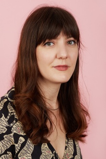 Headshot of artist Cat Willett Jack, wearing a white tigers printed shirt. Backdrop is light pink. Willett's hair is brunette, past the shoulders, with bangs. 