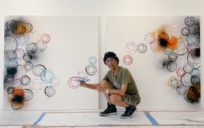 Artist Futura2000 in front of a work of their art, two panels, with circular designs on a white background. 