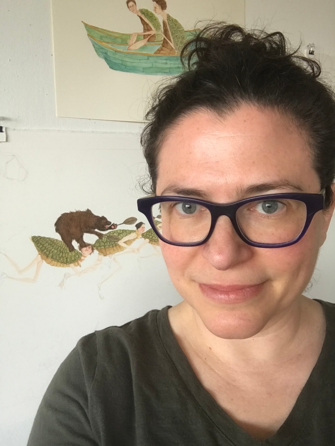 Headshot of Amy Cutler, wearing glasses, and a tshirt, in front of a white wall with illustrations hung in the background. 