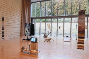 Installation photograph of "350 Million years in the blink of an eye", by Lili Chen, depciting a hardwood floored room, with the far wall being a wall of windows, and various sculputural objects in the foreground. 