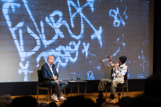 Artist FUTURA2000 (right) in conversation with artist Carlos Mare (left) on stage of the Screening Room, in the Center for the Arts on UB North Campus, as part of the Department of Art's Visiting Artist Speaker Series, on September 25, 2023. The two artists are seated in front of a screen with a projection of Futura's graffiti artist signautre tag, white paint on a black wall. 