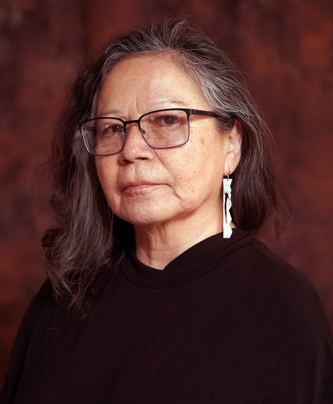 Headshot of Shelley Niro wearing a black garment and beaded earrings, on a burgundy background. 