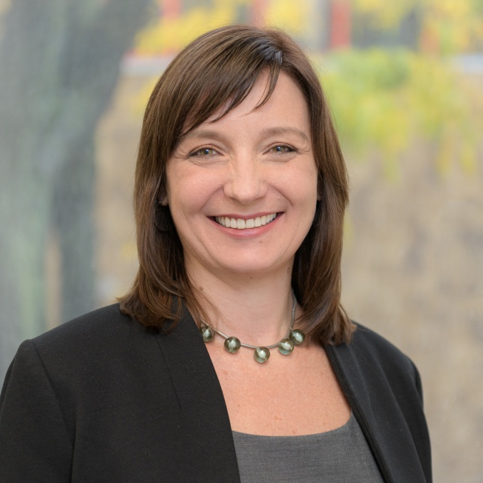 Headshot of Tania Duvergne wearing a gray shirt, black jacket, and out of focus muted colors background. 