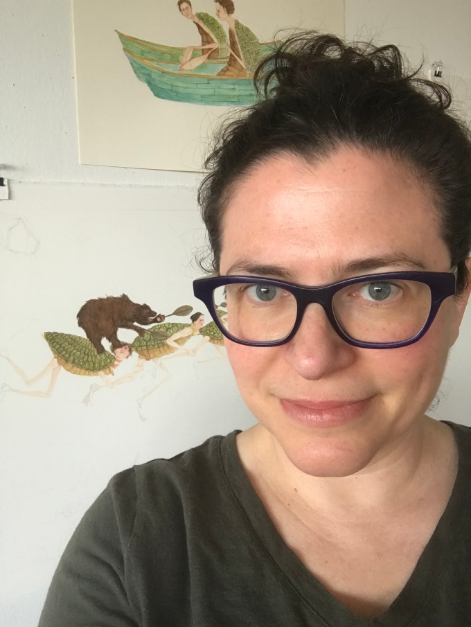 Headshot of Amy Cutler, wearing glasses, and a tshirt, in front of a white wall with illustrations hung in the background.