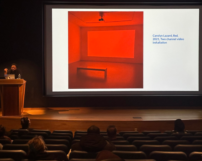 Curator Serubriri Moses, behind a podium on stage of in an auditorium, with a projection screen showing a red lit room and caption "Carolyn Lazard, "Red", 2021, two channel video, installation". 