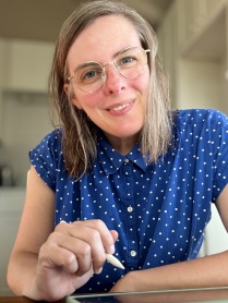 Zoom image: Caitlin Cass headshot wearing a short sleeve blue button up shirt with tiny white polka dots on it, and wearing translucent tan framed glasses, and holding a pen.