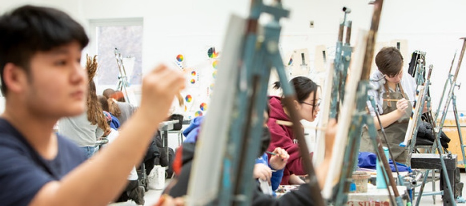 A department classroom, painting studio, a student sits in the foreground of the image, holding a paint brush against a canvas on an eisel, behind him, other students are painting. 