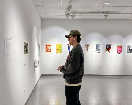 Zoom image: A man wearing a sweatshirt and hat looking at a wall in an art gallery, with penant political posters hanging in the background.