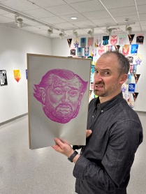 Zoom image: A man in blue button up shirt holding a political cartoon woodcut print of Ted Criz's face.