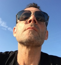 Zoom image: Headhsot of Jonathan Podwil, wearing sunglasses and a black tshirt in front of a blue sky.