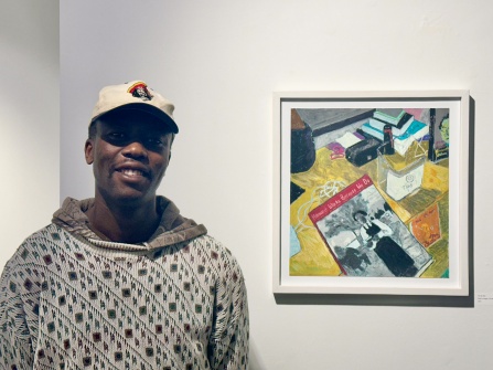 Zoom image: Joel Mulindwa bust up, next to his tip jar oil pastel drawing, in the Project Space gallery, for exhibition "Impressions of Gratuity".