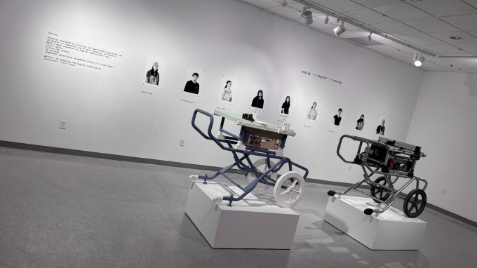 Zoom image: two contractor table saws on pedestals in the middle of an art gallery, with exhibition text and artist headshots hung on the rear wall.