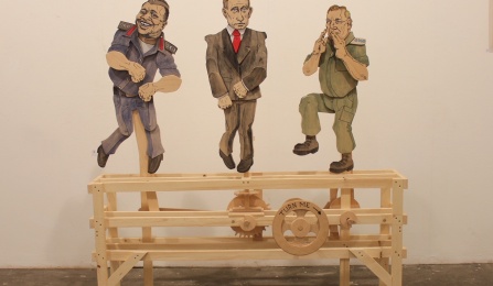 Wooden sclupture by Frederick Wright Jones, depicting 3 world leader caricatures on top of a wooden mechanical base. 