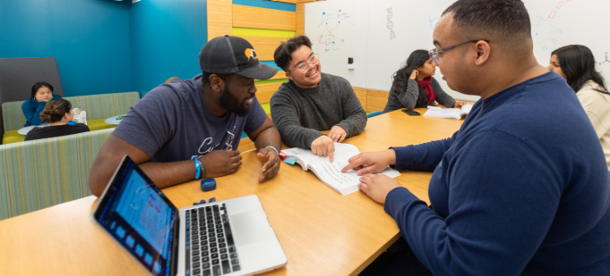 A small group of students sit around a table in a brightly colored study room, discussing a textbook together. One student points to a page while others listen and smile, with a laptop open on the table and additional students studying in the background. 
