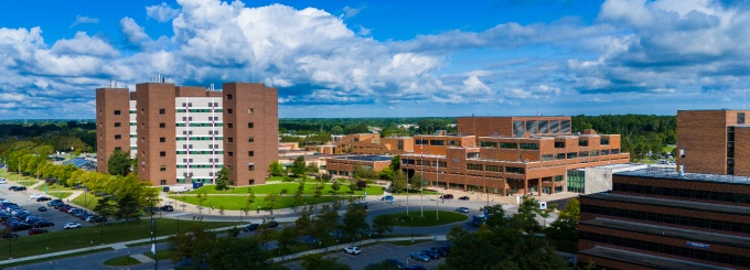 An aerial view of the University at Buffalo&rsquo;s North Campus shows multiple brick academic and residential buildings surrounded by green lawns, roads, and parking areas under a bright blue sky with scattered clouds. 