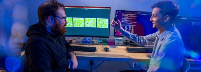 Two researchers sit at a desk in a blue-lit lab or office, discussing data while looking at computer monitors displaying colorful grid-based visualizations and lines of code; one holds up a smartphone to show the other something on the screen. 