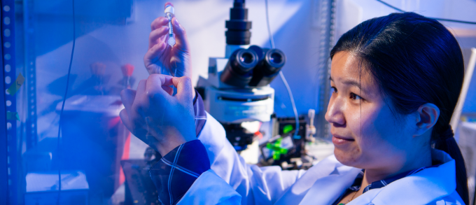 A researcher in a white lab coat examines a small sample tube while working at a lab bench illuminated in blue light, with a microscope and scientific equipment visible behind her. 