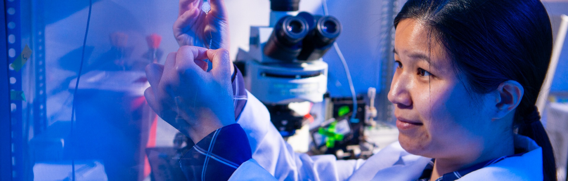 A student researcher in a lab coat examines a small sample tube under blue light while standing beside a microscope in a biology lab. 