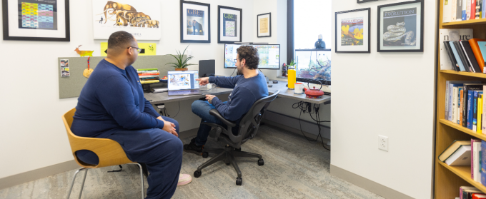 Two researchers sit in a genomics lab office, reviewing data on a laptop and desktop monitors, with framed scientific journal covers and biology references on the walls as they discuss genomic analysis results. 