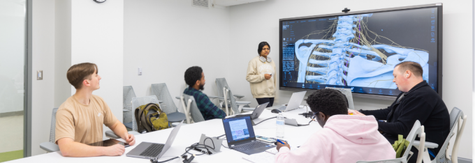 Students sit around a conference table with laptops while an instructor presents a 3D anatomical visualization of the human torso and nervous system on a large digital display during a cellular signaling class. 