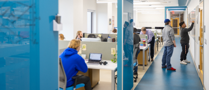 Students and staff work and collaborate in a modern academic office space with open cubicles and glass-walled corridors. Some people sit at desks using computers, while others stand in the hallway discussing and reviewing materials posted on the wall. 