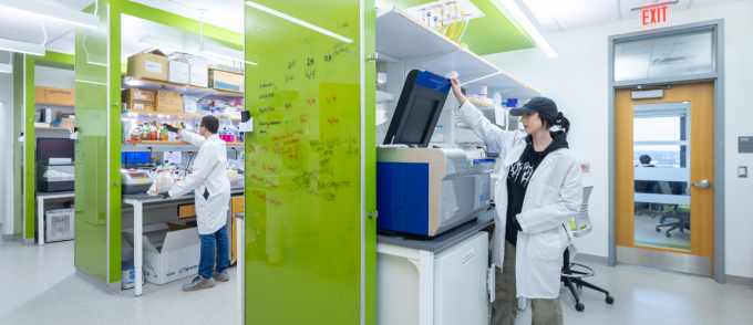 Student researchers work in a modern laboratory with bright green glass partitions. One researcher in a lab coat operates a large scientific instrument, while another prepares samples at a lab bench surrounded by shelves of supplies and equipment. 