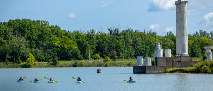 Students kayak across a calm lake with bright green paddles, passing a set of tall stone columns along the shoreline on a sunny day. 