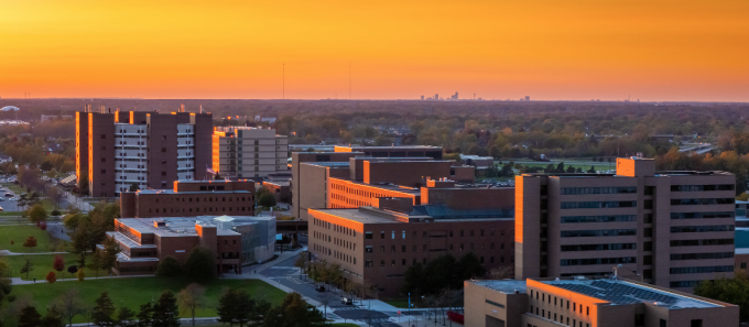 An aerial view of the University at Buffalo&rsquo;s North Campus at sunset, with academic buildings and residence halls bathed in warm golden light, surrounded by tree-lined green spaces and the distant city skyline on the horizon. 