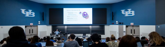 A wide view of a large University at Buffalo lecture hall shows a professor presenting at the front of the room beneath a large screen displaying a biology lecture slide about the cell nucleus. Students sit in tiered seating, many using laptops, with the UB logo visible on the walls. 