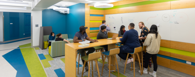 Students collaborate in a bright, modern study space with a long table and whiteboard walls. Several students sit and talk around the table with laptops and books, while others write and discuss ideas at the whiteboard. Additional students work in small seating areas nearby. 