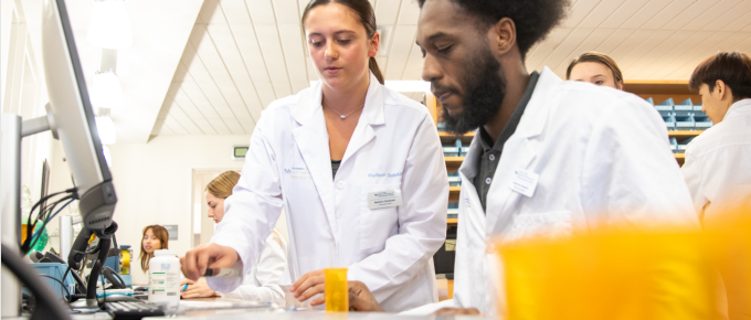 Students wearing lab coats work together at a laboratory bench, measuring and handling materials during a hands-on science activity. One student demonstrates a step while others observe and assist in the background.