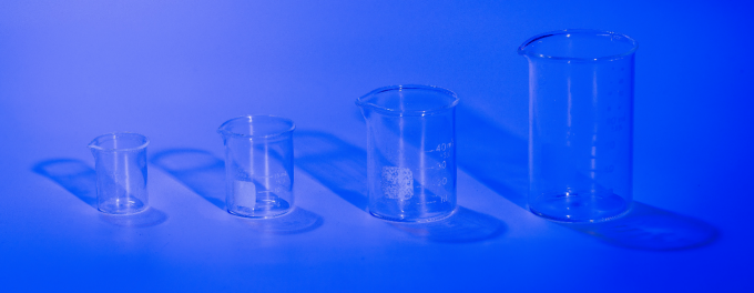 Four empty laboratory beakers of increasing size are arranged in a row on a flat surface, lit with blue lighting that casts soft shadows behind them.