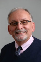 Prof. Joseph Gardella wearing a dark v-neck sweater, blue tie, a pink collared shirt and dark framed glasses. 