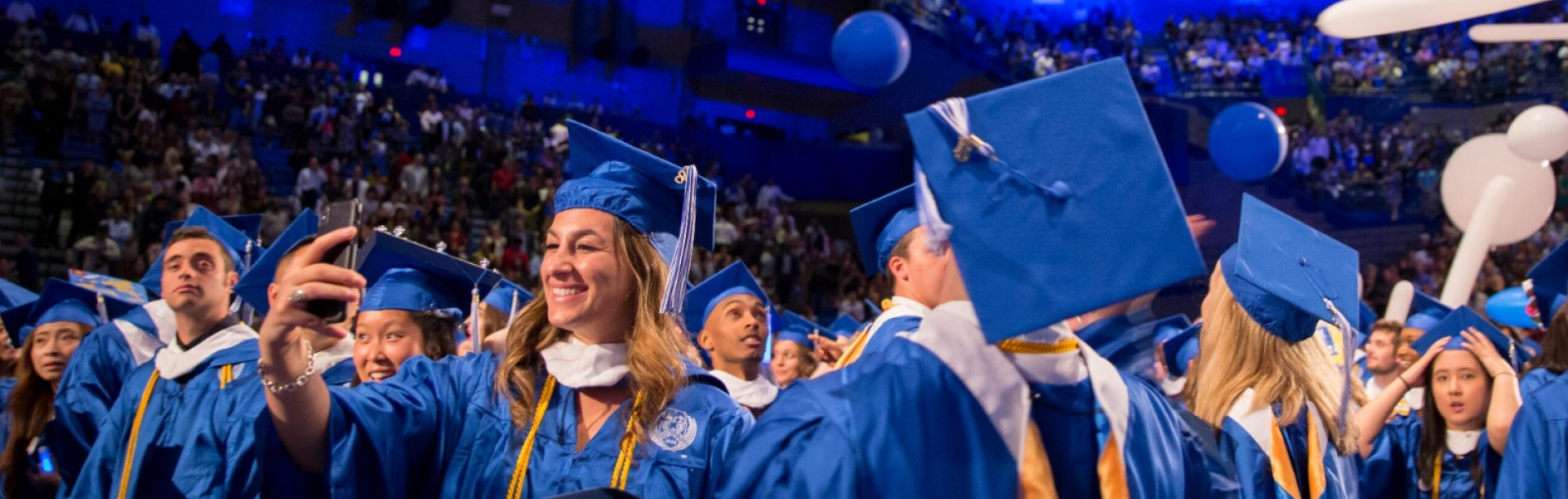 Students in commencement cap and gowns celebrate in Alumni Arena, with blue and white balloons dropping from the ceiling. 
