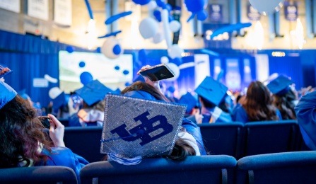 A student with their back to the camera wearing a cap and gown takes a photo. 