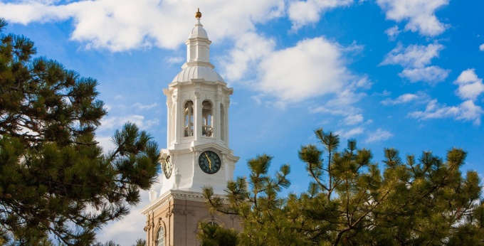 Pictured, UB’s iconic South Campus clock tower atop Edmund B. Hayes Hall. A stately white dome with ornate trim and openings that partially expose the large bells (known as the Westminster chimes) housed inside the tower, framed by a vibrant blue sky with white clouds and long-needled pines. The center portion of the building beneath this clock tower, now Hayes Hall, was built in 1874 to house the Insane Department of the Erie County Almshouse. It eventually expanded into a 400-bed hospital before being converted for academic use in 1928. 